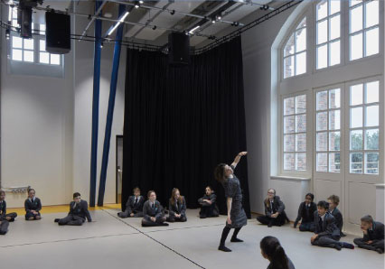 Interior of a studio space with tall arched windows, a wooden floor and dark drapes, young people perform in the space