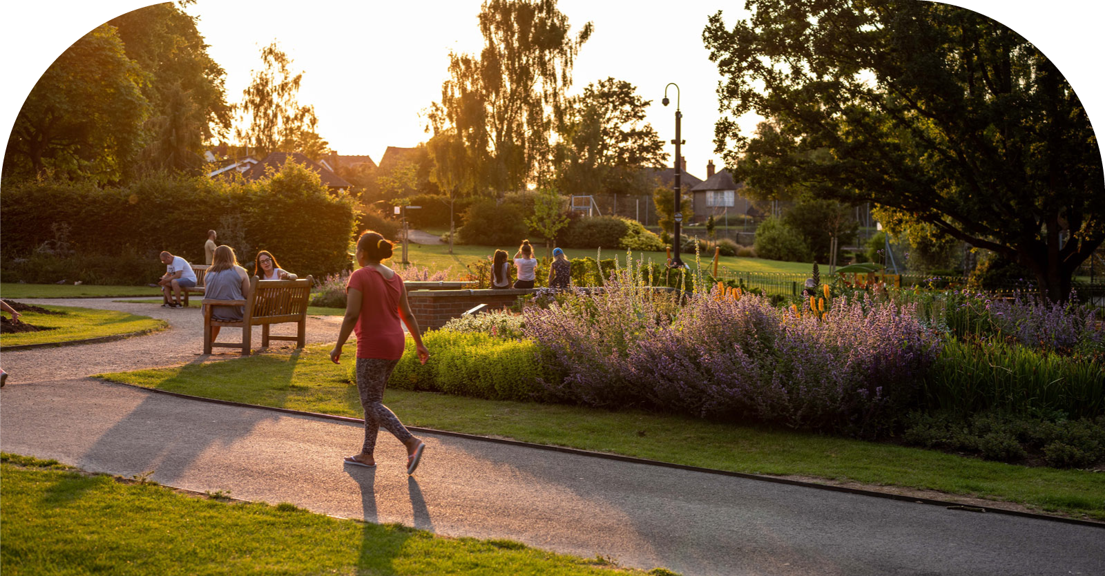 Person in a red top walking through a park at golden hour among trees and flowers
