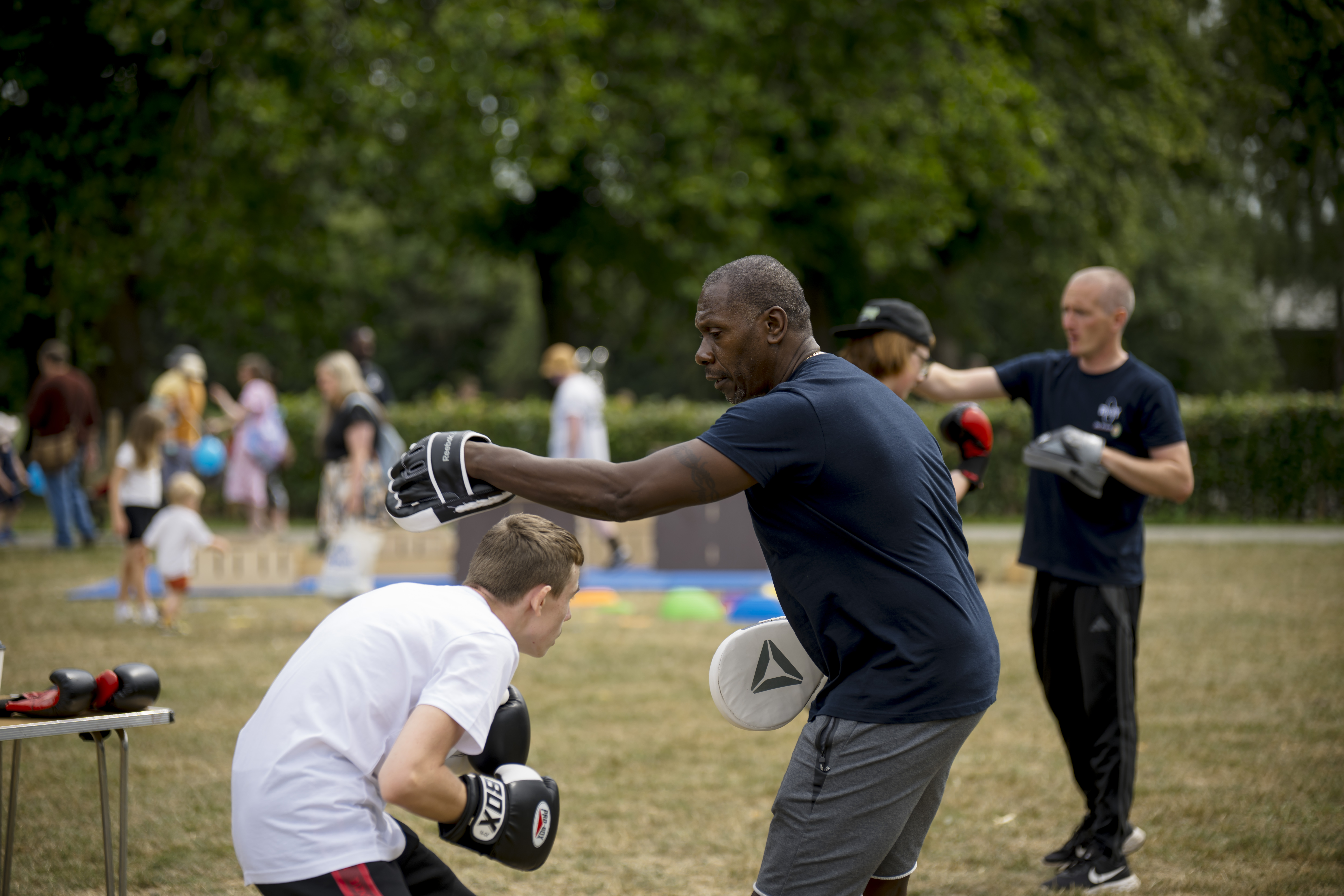 Child in boxing gloves punching focus pads held by a coach at an outdoor family activity day