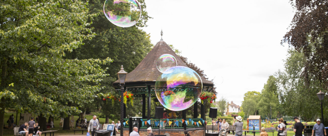 Caldecott park in the Summer during an event, with the bandstand in the background, groups of people enjoying themselves and bubbles blowing in front of the bandstand