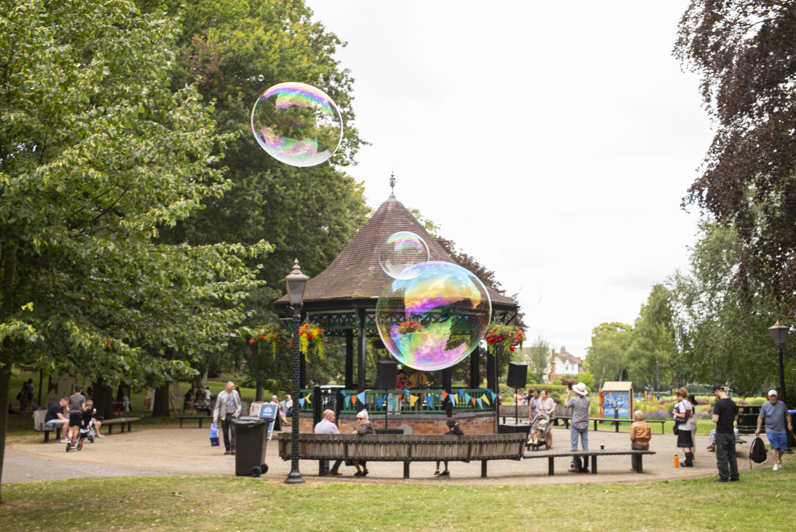 Caldecott park in the Summer during an event, with the bandstand in the background, groups of people enjoying themselves and bubbles blowing in front of the bandstand