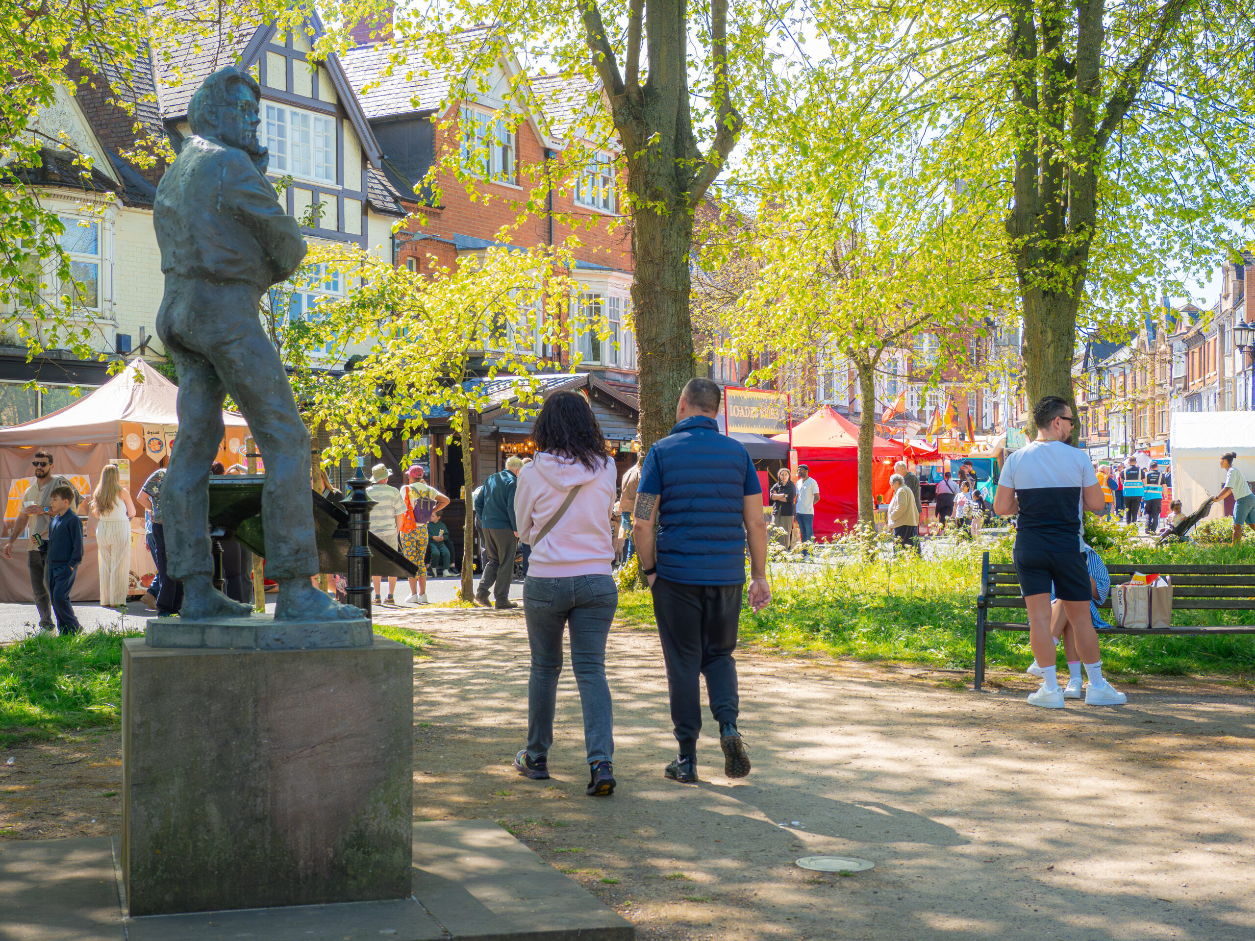 Crowds browsing market stalls at an outdoor food festival in the park, beside the bronze rugby player statue