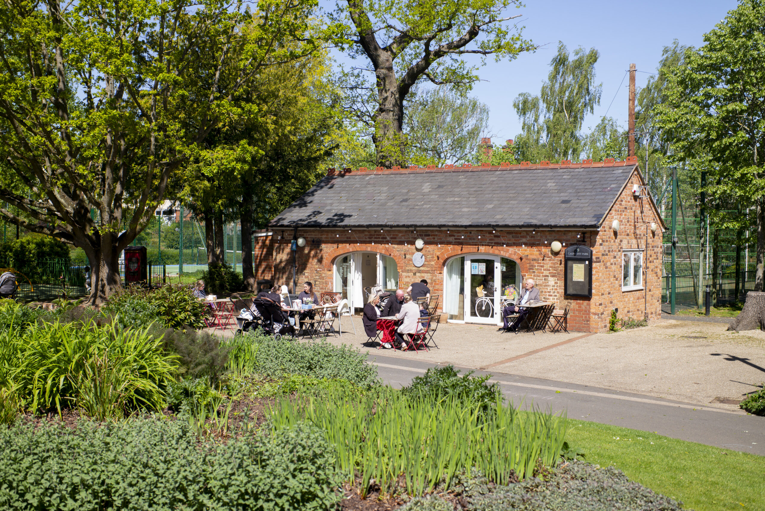 Brick park pavilion cafe with people seated outside among flower beds and lawns