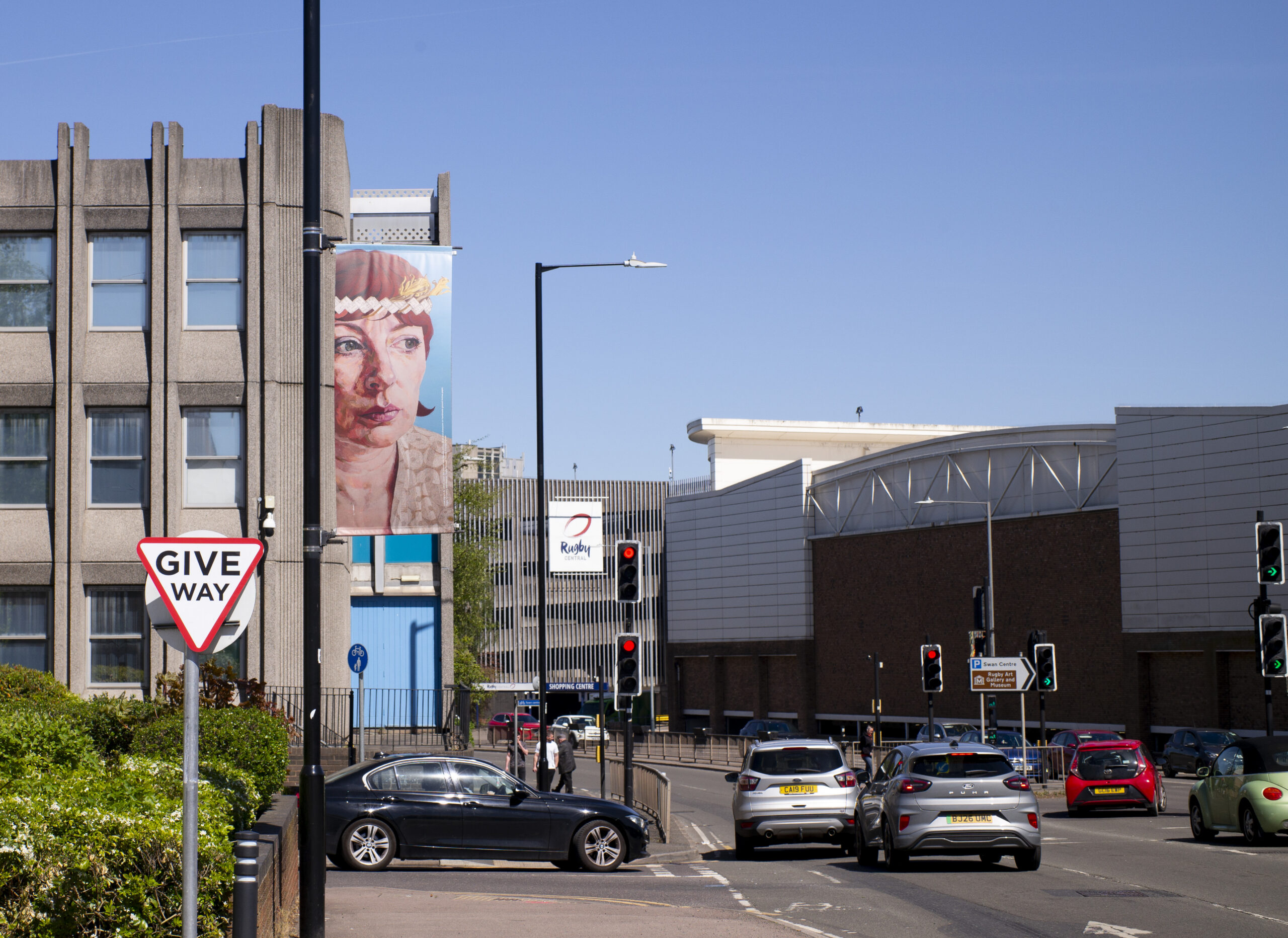 A busy town-centre street with a large painted mural of a face on a lamp post banner and traffic passing