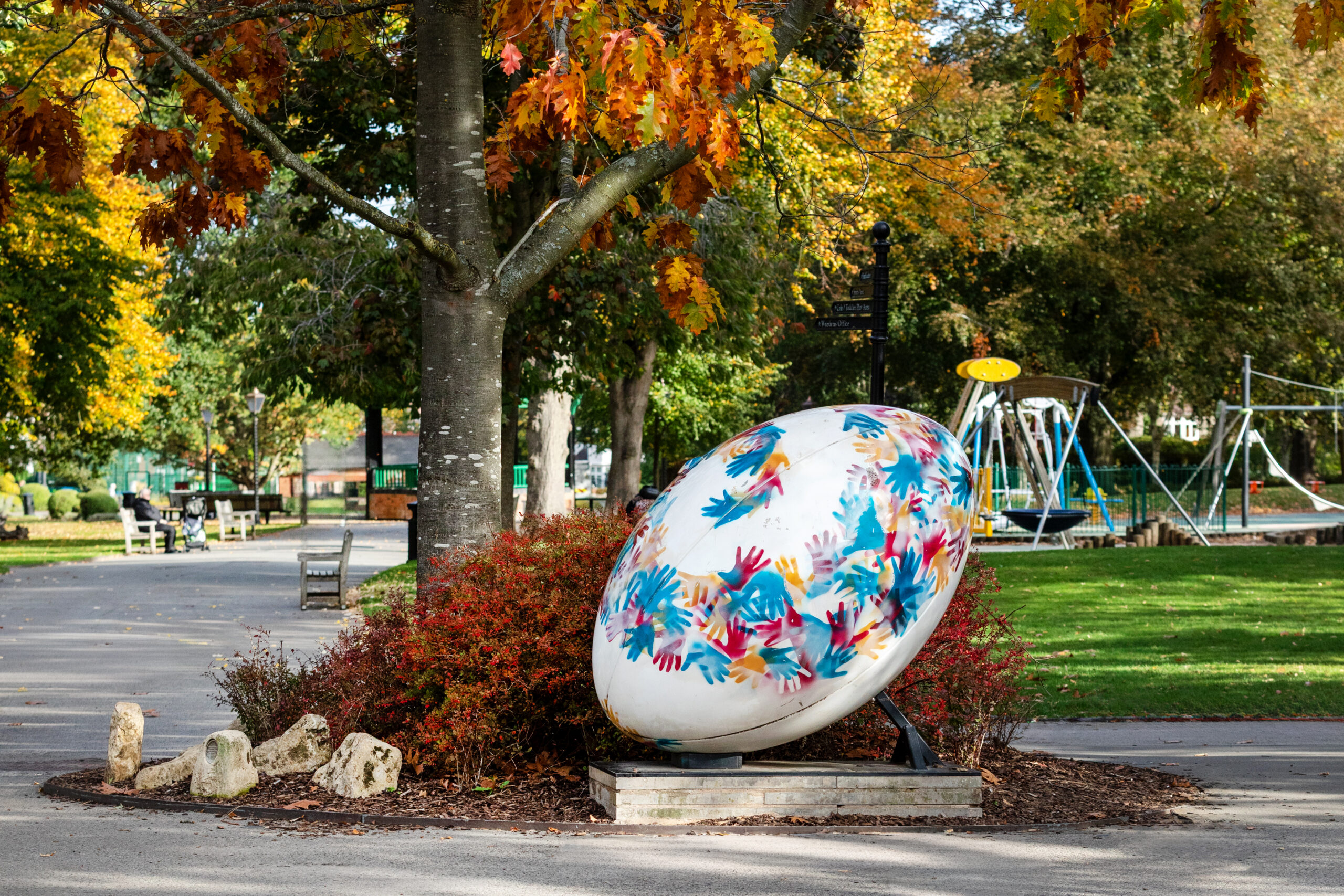 Large decorated rugby ball sculpture painted with colourful handprints on a park path in autumn