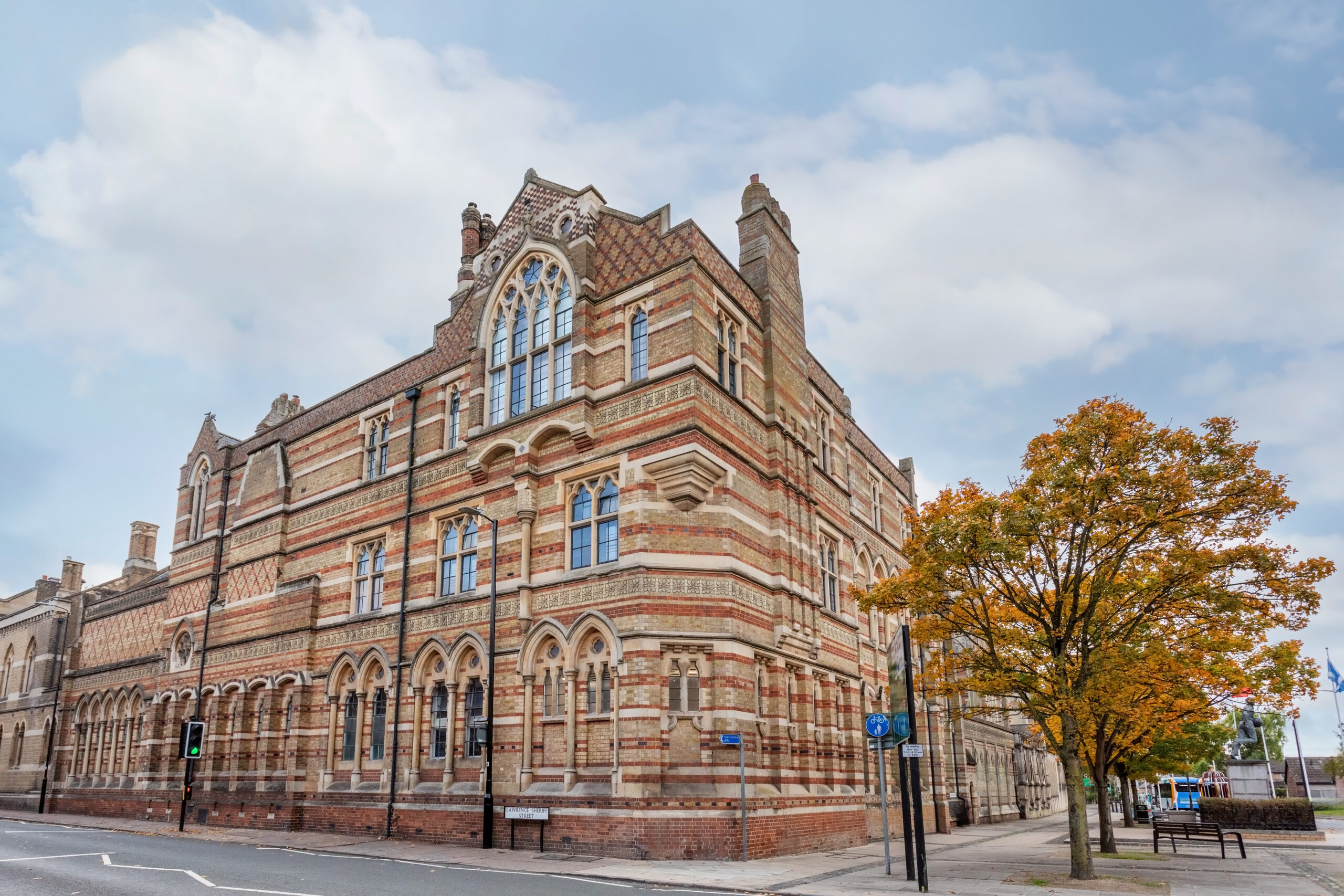 Ornate Rugby School building on a street corner beside an autumn-coloured tree.