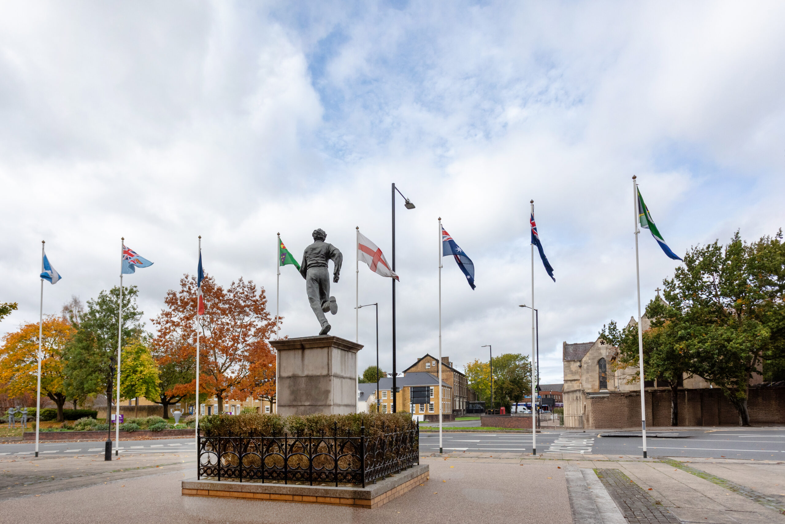 Bronze statue of a Victorian rugby player on a plinth with flagpoles under a cloudy sky