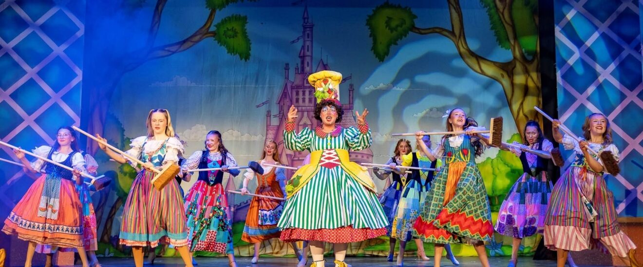 Pantomime dame in costume with a dancing ensemble cast in colourful costume.