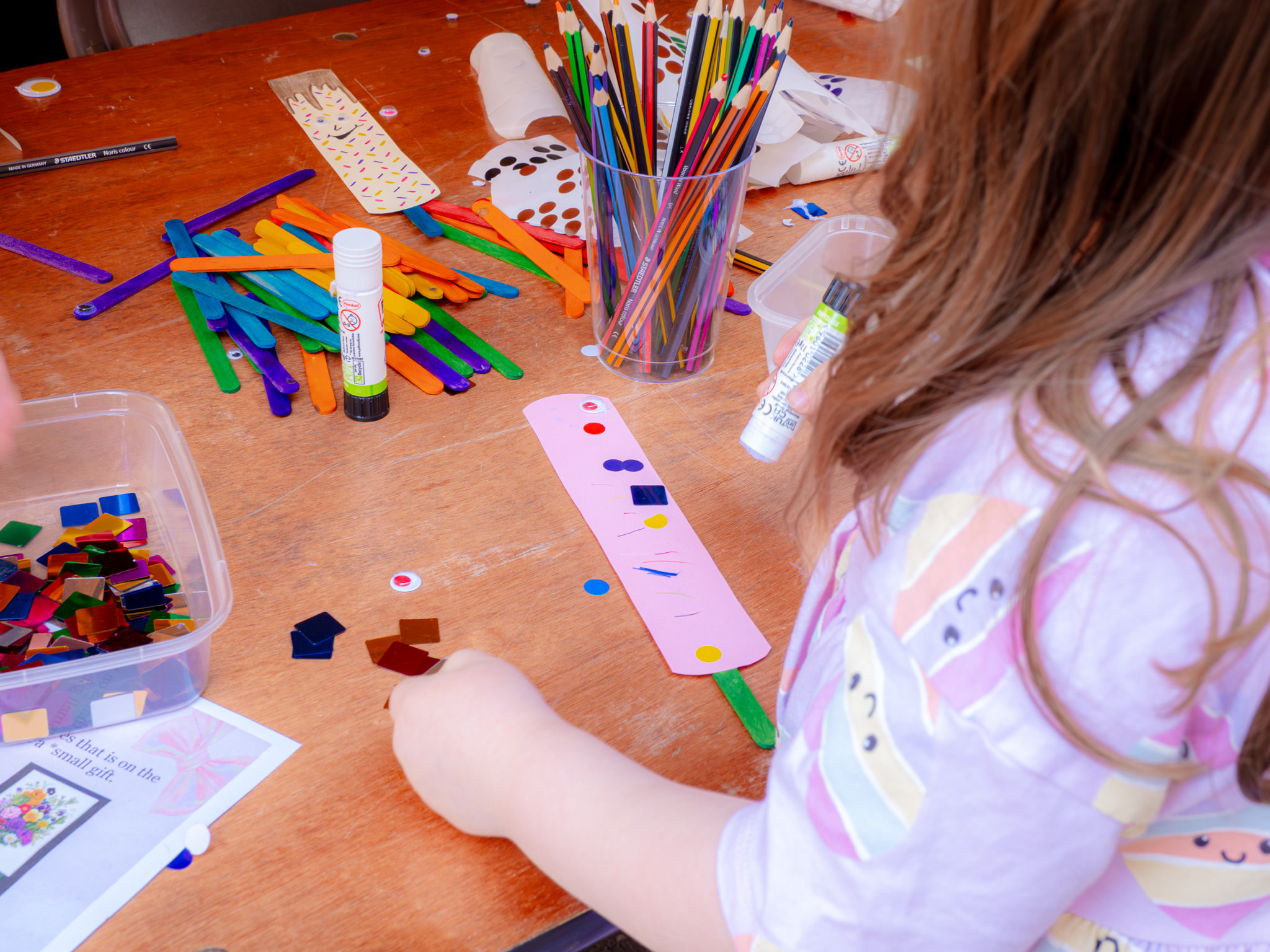 table with craft supplies on it and a child gluing down colourful shapes.
