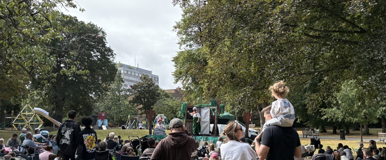 A crowd of all ages gathered in Caldecott Park. People are sat on camping chairs or the ground watching a performance on a pop up stage. A child is on it's parents' shoulders in the foreground, the sun is shining and the trees in the background are in full leaf.