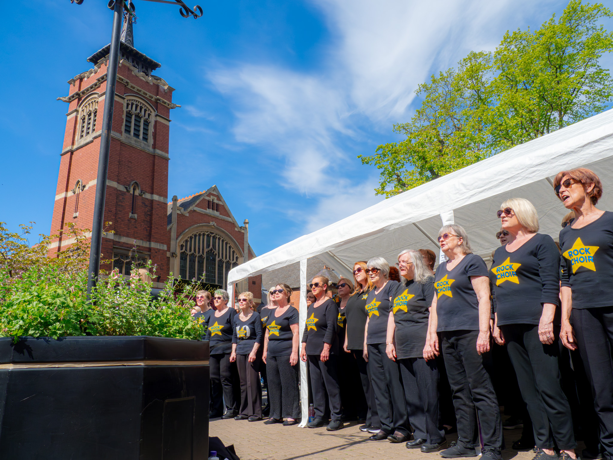 A rock choir performing at an outdoor event under a marquee and in front of a church building, with a bright blue sky in the background