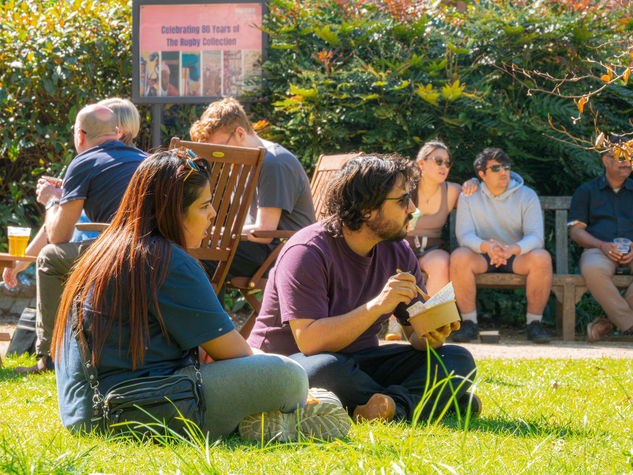 A group of people sat on the grass together eating and drinking at an outdoor event