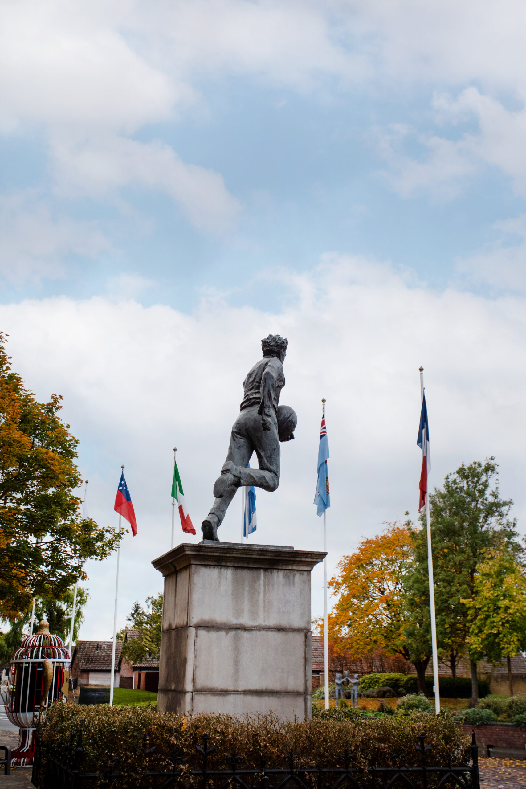 Statue of a rugby player running with a ball, on a stone plinth with flagpoles behind against a blue sky