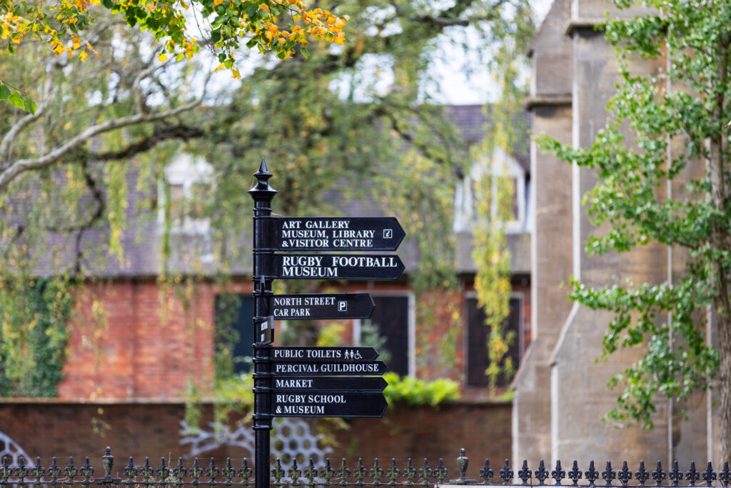 Black cast-iron direction signpost with St Andrews Church Gardens in the background, the honeycomb sculpture is visible and the trees are in autumn colours. The directional sign post is directing you to the left towards the town centre.