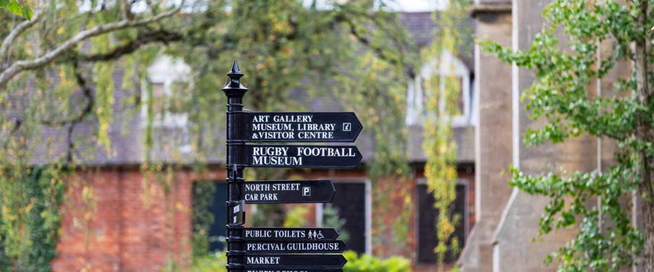 Black cast-iron direction signpost with St Andrews Church Gardens in the background, the honeycomb sculpture is visible and the trees are in autumn colours. The directional sign post is directing you to the left towards the town centre.