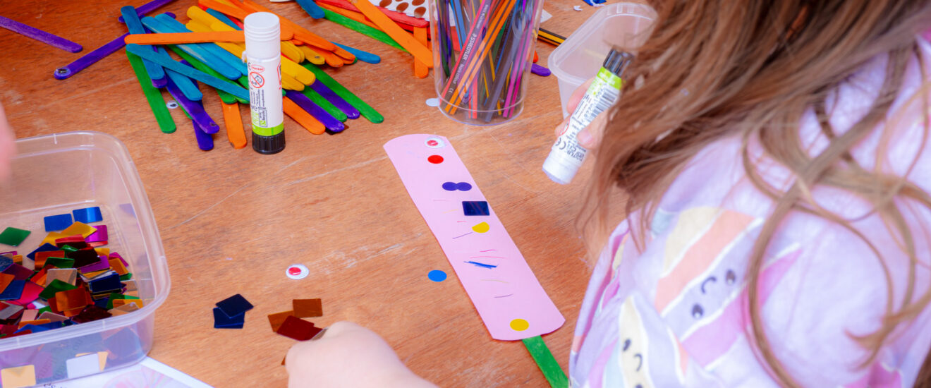 table with craft supplies on it and a child gluing down colourful shapes.